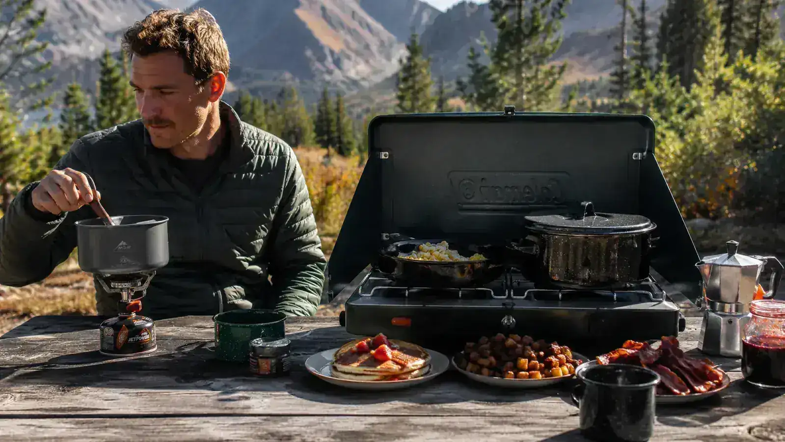 Group of four campers eating hot food around a camp table at dusk, plates full, two-burner stove visible in the background