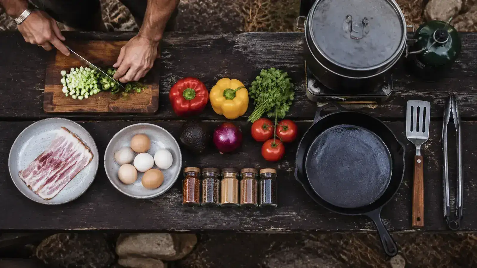 Campsite meal prep station with cutting board, knives, spices, and a two-burner stove in the background